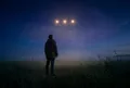 低角度仰拍一個人在開闊原野上抬頭仰望夜空，三顆柔和金色光球在上方排成一直線，深靛藍與紫色夜空與薄霧，暖光氛圍安定耐心
