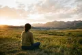 女性背影坐於空曠草地邊緣，身後是廣闊天空與遠山，傍晚金光從左側低角度灑落，人物佔畫面左下角三分之一