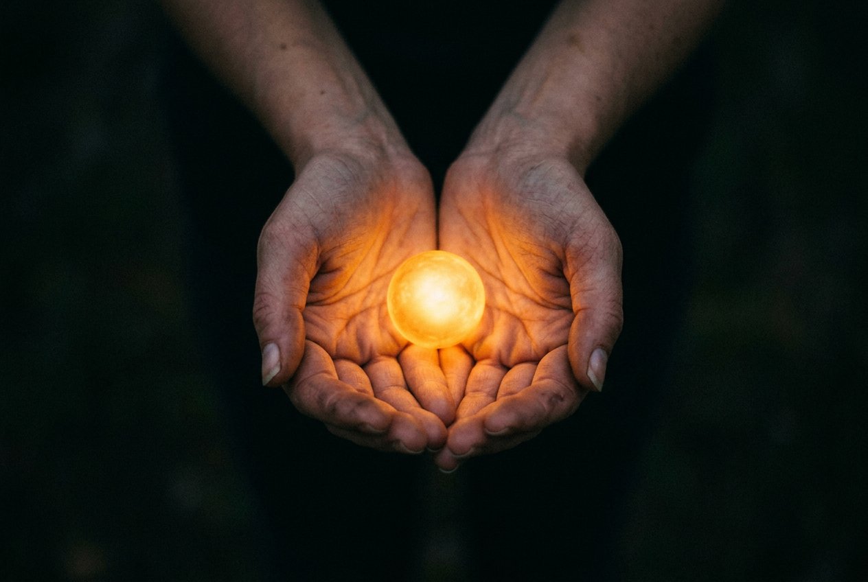 雙手捧住一顆發光的小光球，從略高角度拍攝，雙手位於畫面下方居中，周圍黑暗向邊緣消退，溫暖金光映照在皮膚上