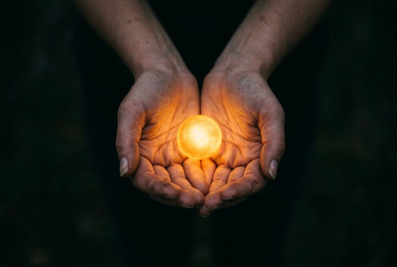 雙手捧住一顆發光的小光球，從略高角度拍攝，雙手位於畫面下方居中，周圍黑暗向邊緣消退，溫暖金光映照在皮膚上