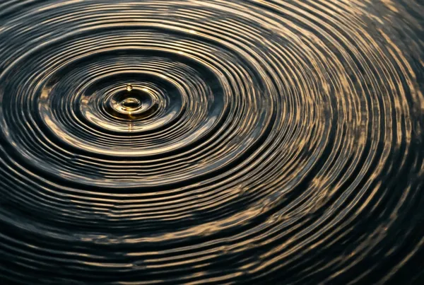 俯拍水面漣漪從單一水滴衝擊點向外擴散，同心圓充滿整個畫面，傍晚斜光在水面形成明亮高光與暗色波谷，衝擊點位於左上三分之一處